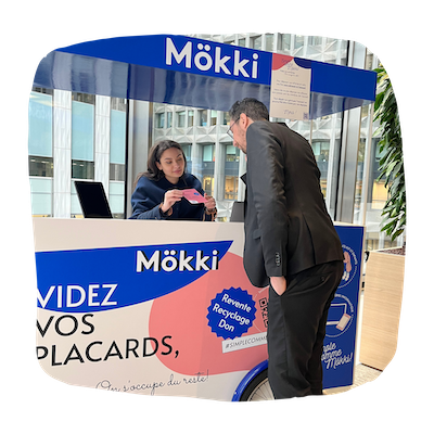 Un kiosque de l'entreprise Mökki est installé dans le hall d'un bureau. Une personne de Mökki explique le fonctionnement de la collecte des vêtements à un employé des bureaux.