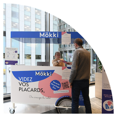 A Mökki kiosk is installed in the lobby of an office building. A Mökki staff member hands a flyer to an employee.